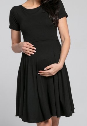 Kobieta w ciąży w czarnej sukience z krótkimi rękawami, delikatnie trzymająca oba rękami ciążowy brzuszek na jednolitym szarym tle.