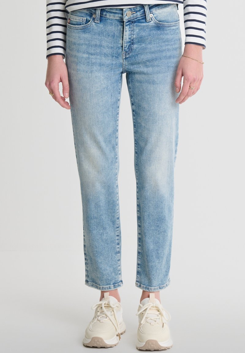 Gai zilu džinsi ar taisnu piegriezumu, ar nelielu nolietojumu un rullētām malām. Apvienoti ar krēmkrāsas kedām un svītrainu augšdaļu.