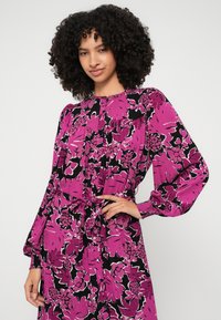 Cvetna obleka z drznimi roza in črnimi vzorci, dolgimi rokavi, okroglim izrezom in privezanim pasom za prilagojeno silhueto.