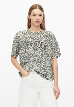 Femme aux cheveux raides portant un T-shirt gris et noir imprimé léopard avec le texte "UNIQUE" et un pantalon blanc, une main dans la poche.
