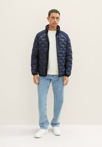 Giubbotto imbottito blu navy con chiusura a zip, dotato di colletto alto e polsini elastici. Indossato sopra un maglione leggero e abbinato a jeans chiari.