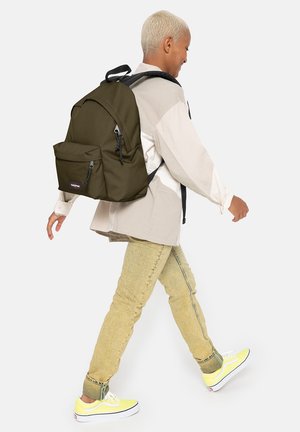 Mochila de color verde oliva con un bolsillo frontal y cremalleras negras, llevada por una persona con una camisa clara y pantalones beige, caminando con zapatos amarillos.