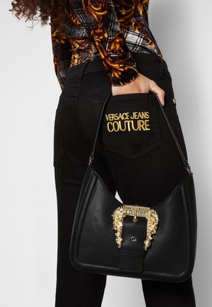 Femme portant un jean noir avec une broderie dorée "Versace Jeans Couture" sur la poche, tenant un sac à main en cuir noir avec une boucle dorée ornée.