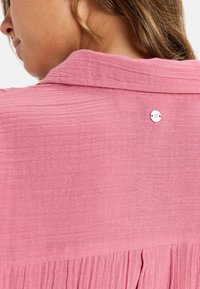 Rosa, strukturiertes Stoffoberteil mit Kragen, das über eine Knopfdetaillierung im Rücken verfügt. Das Design umfasst gefaltete Akzente entlang des Saums.