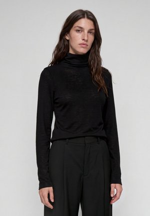 Donna con lunghi capelli castani che indossa un maglione nero a collo alto e pantaloni neri, in piedi contro uno sfondo chiaro e semplice.