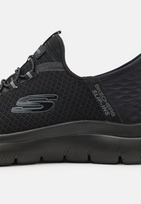Black Skechers slip-on shoes with mesh upper, textured accents, thick cushioned sole, and logo detail on the side.