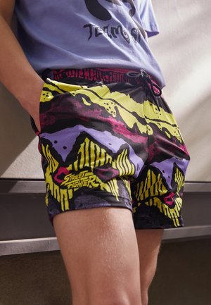 Osoba nosząca kolorowe szorty z motywem Street Fightera w abstrakcyjne wzory w żółtym, fioletowym i różowym kolorze, stojąca z jedną ręką w kieszeni.