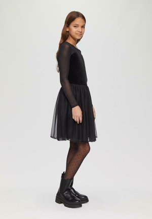 Robe noire avec des manches longues en voile, un haut ajusté et une jupe en tulle. Portée avec des collants noirs à pois et des bottes de cheville noires épaisses.
