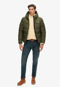 Mandlig model iført en grøn hættetrøje-pufferjakke, cremefarvet kabelstrikket sweater, blå jeans, brun læderbælte og brune snørebåndsstøvler, stående.
