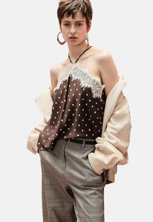 Giovane che indossa un top a pois con scollo all'americana e rifinitura in pizzo, giacca beige oversize caduta sulle spalle e pantaloni a quadri con le mani in tasca.