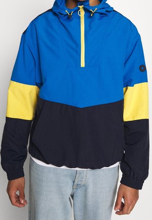 Person, die eine blau-gelb-schwarz colorblock Kapuzenpulloverjacke mit gelbem Frontreißverschluss und elastischen Bündchen trägt.