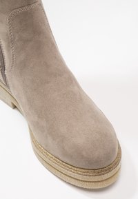 Bottine en daim marron clair avec bout arrondi, fermeture éclair latérale et semelle en caoutchouc beige texturée sur fond blanc.