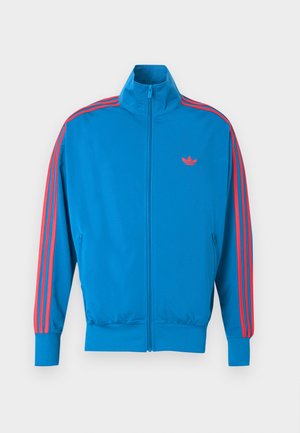 Blue athletic jacket made from smooth fabric. Features red stripe accents along the sleeves and a small logo on the chest. Zipper front closure.