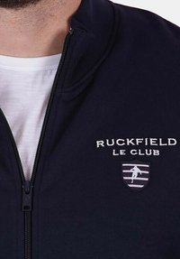 Homme portant une veste zippée bleu marine sur une chemise blanche, avec "Ruckfield Le Club" et un emblème sportif sur la poitrine gauche.