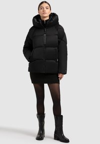 Naistev naine, seljas must puffer-jakk suure kapuutsiga, must mini-seelik, läbipaistvad sukkpüksid ja mustad keskmise pikkusega nahk-kingad, seistes tasase tausta ees.