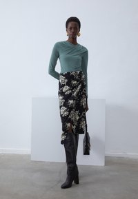 Haut vert clair à manches longues avec des détails froncés, associé à une jupe portefeuille noire à motifs floraux, des bottes noires arrivant jusqu'aux cuisses et un petit sac noir.