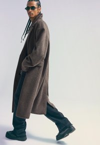 Hnedý, dlhý vlnený kabát s voľným strihom; skombinovaný s čiernymi širokými nohavicami a masívnymi čiernymi topánkami. Doplnkovaný okrúhlymi, tmavými slnečnými okuliarmi.