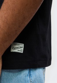 Κοντινό πλάνο του κορμού ενός ατόμου που φοράει μαύρο πουκάμισο με λευκή ετικέτα και ανοιχτόχρωμο μπλε τζιν παντελόνι.