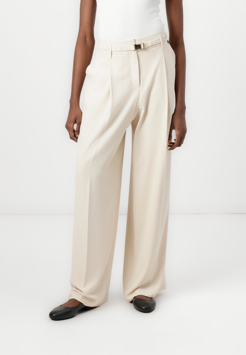 Beige weite Hosen aus glattem Stoff, mit einem Taillengürtel und gefaltetem Vorderteil. Kombiniert mit schwarzen flachen Schuhen.