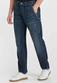 Jeans in denim blu scuro con design a gamba dritta, dotati di dettagli cuciti e tasche laterali, abbinati a sneakers bianche.