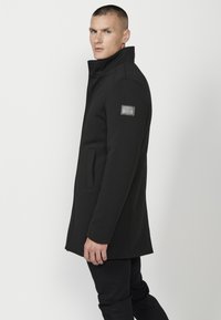 Cappotto in misto lana nera con colletto strutturato, tasche laterali e una toppa sul maniche sinistro. Il design è sartoriale e aderente.