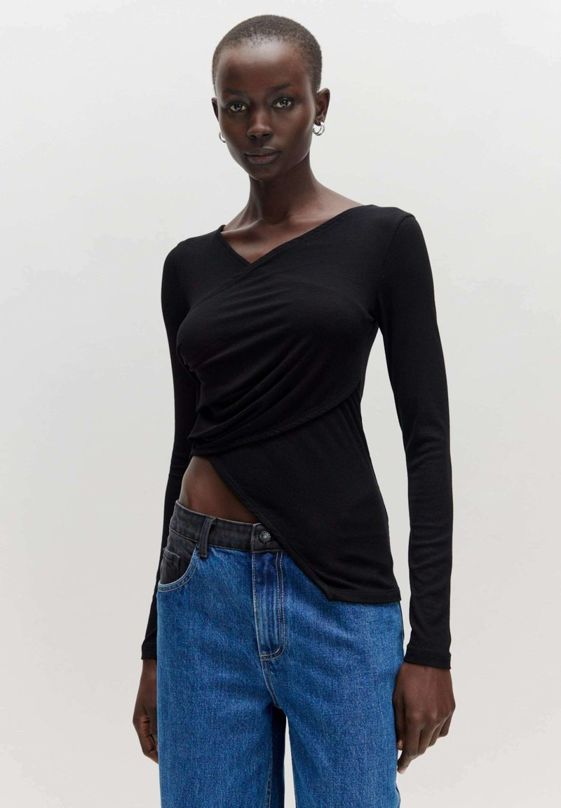 Schwarzes Langarmoberteil mit Wickeldesign, V-Ausschnitt und strukturiertem Stoff, kombiniert mit mittelhohen blauen Jeans.