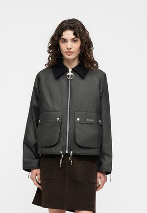 Göndör hajú nő fekete cipzáros dzsekit nagy első zsebekkel és sötétbarna szoknyát visel, tiszta világos háttér előtt áll.