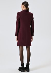 Cappotto in misto lana color bordeaux con silhouette sartoriale, maniche lunghe, colletto a revers e dettagli della cucitura sul retro, abbinato a stivali alla caviglia neri.