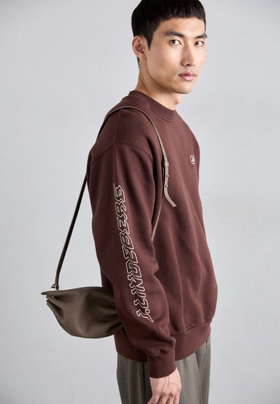 Καφέ φούτερ με λογότυπο στο αριστερό μανίκι, συνδυασμένο με μια μαλακή καφέ τσάντα ώμου με καμπυλωτό σχέδιο και ρυθμιζόμενο λουράκι.