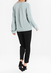 Blouse à manches longues bleu clair à motifs avec une coupe décontractée, associée à un pantalon noir fuselé et des chaussures à talons noires.