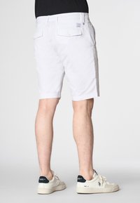 Pantaloni corti bianchi in cotone con tasche, di fronte piatto e orlo dritto. Indossati con sneaker crema e blu navy. Design semplice e pulito.