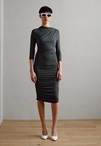 Temno zelena obleka s dolgimi rokavi in nabranim teksturo, v kombinaciji z belimi visokimi petami in oversize belimi sončnimi očali.