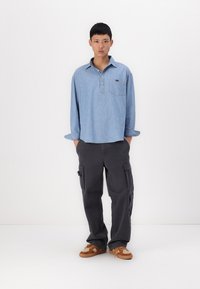 Cămașă albastră deschis cu mâneci lungi, asociată cu pantaloni cargo gri închis cu buzunare laterale și pantofi sport maro cu tălpi albe.