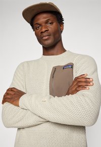 Homme portant un pull texturé beige avec une poche zippée marron sur la poitrine et une casquette marron, les bras croisés, regardant la caméra avec confiance.