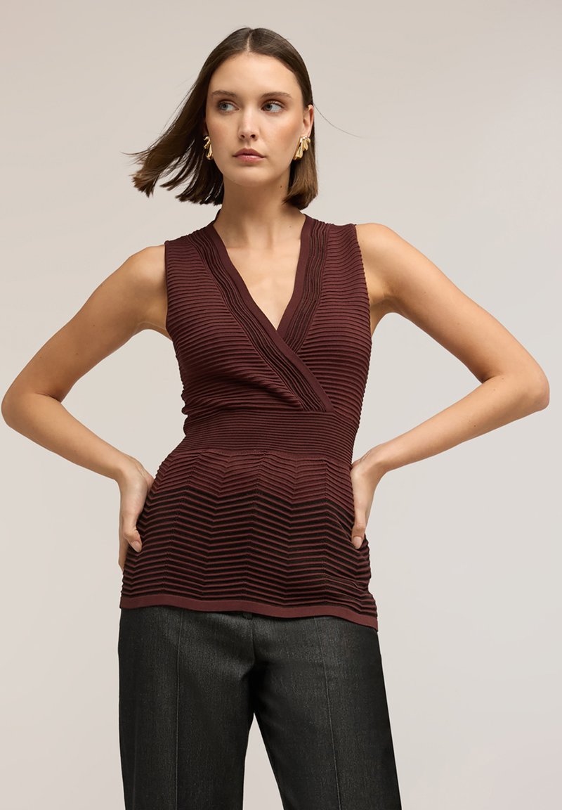 Mujer con cabello corto castaño, que lleva pendientes dorados, top sin mangas texturizado de color burdeos y pantalones negros, con las manos en las caderas, mirando de lado.