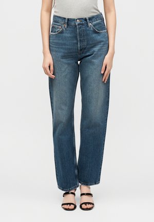 Frau trägt hochtaillierte, gerade geschnittene blaue Jeans kombiniert mit schwarzen offenen Sandalen und einem hellgrauen ärmellosen Oberteil.