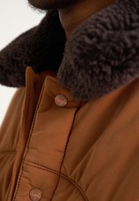 Casaco acolchoado castanho com gola de pelo escuro. Apresenta botões de tom cobre e textura suave do tecido.