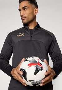 Chaqueta deportiva negra con cremallera y un logo dorado de "KING" junto al emblema de Puma. Se sostiene un balón de fútbol blanco con patrones en negro y gris y un logo rojo de Puma.