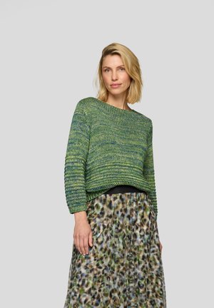 Grüner Strickpullover mit strukturiertem Muster, weitem Ausschnitt und schmal zulaufenden Ärmeln, kombiniert mit einem gemusterten, fließenden Rock in Grün- und Brauntönen.