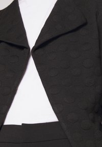 Veste noire texturée avec des motifs circulaires en relief portée sur une chemise blanche unie et associée à un pantalon noir.