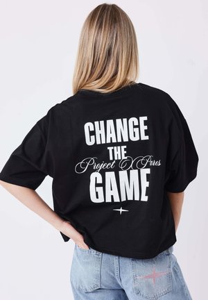 Persona con capelli biondi indossa una maglietta nera con testo bianco "CHANGE THE Project X Paris GAME" e jeans azzurri con una stella rosa sulla tasca.