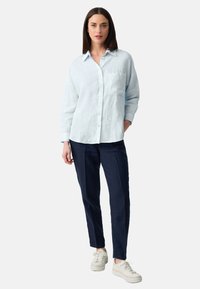 Vrouw die staat en een lichtblauw button-up overhemd met lange mouwen, donkerblauwe broek en witte sneakers draagt, op een effen grijze achtergrond.