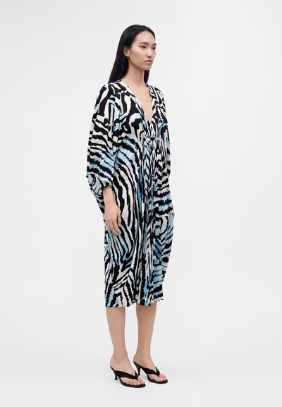 Γυναίκα που φοράει ένα φαρδύ φόρεμα μήκους μέχρι το γόνατο με ριγέ μοτίβο ανάλογο ζέβρας σε μαύρο, άσπρο και μπλε και μαύρα σανδάλια με τακούνι, στέκεται μπροστά σε απλό φόντο.