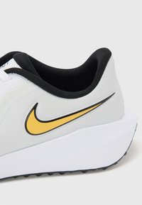 Weißer Sportschuh mit schwarzem Kragen, der eine gelbe Nike-Swoosh-Grafik aufweist. Strukturiertes Material mit einer dicken weißen Sohle und schwarzem Profil.