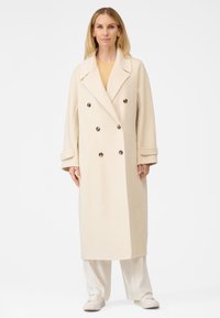 Beige dobbeltbrystet frakke med store klapper, med mørke knapper, skræddersyet pasform og en glat tekstur, båret over lyse bukser.
