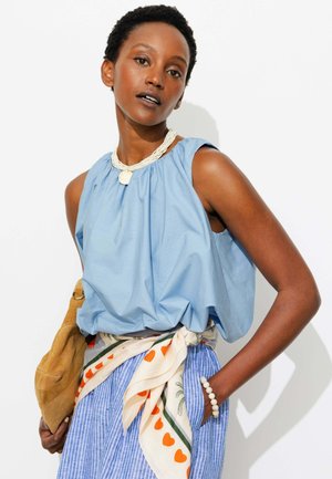 Femme portant un haut bleu sans manches, une jupe rayée avec une ceinture foulard à motif de cœurs, un collier et un bracelet de perles blanches, tenant un sac à main camel.