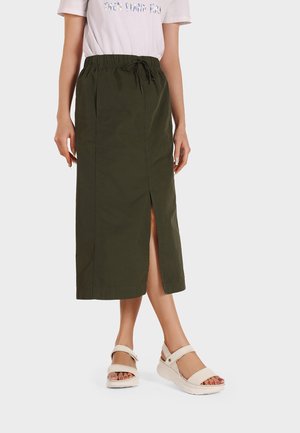 Donna che indossa una gonna midi verde oliva con spacco frontale, t-shirt bianca con grafica e sandali beige con cinturini alla caviglia.