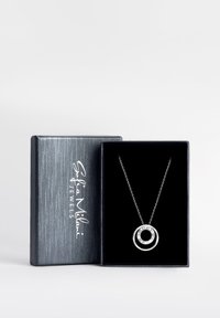 Silverhalsband med en cirkulär berlock som har en central svart cirkel omgiven av klara ädelstenar, presenterad i en grå låda med en texturerad yta.