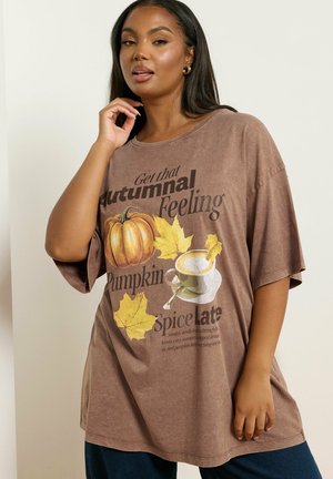 Kobieta nosząca brązowy, oversize t-shirt z jesiennymi liśćmi, dynią i filiżanką latte z tekstem o jesieni i przyprawach do dyni.