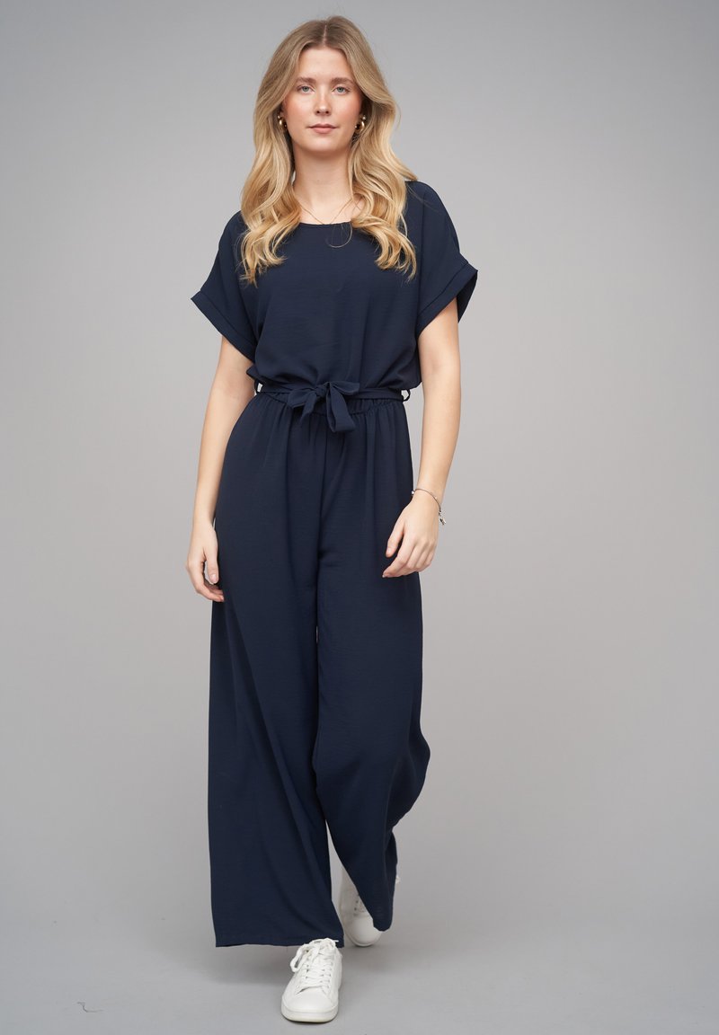 Femme blonde portant une combinaison bleu marine avec une taille nouée, des manches retroussées et des baskets blanches, debout devant un fond gris.
